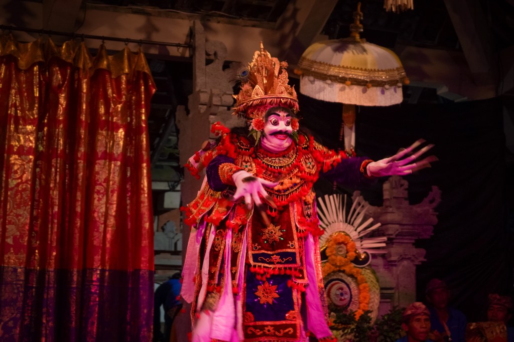 **Title:** Majestic Jauk Manis Dance – The King of Giants in Balinese Tradition **Alt Text:** A Balinese performer in the Jauk Manis dance, dressed as the king of giants with a red ornate costume, golden crown, long painted nails, and a striking mask, performing on stage with traditional temple decorations in the background.