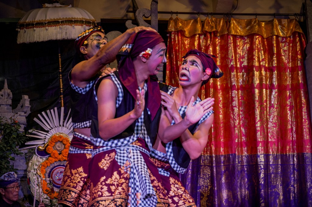 Three Balinese performers in traditional clown costumes and expressive makeup act out a comedic scene on stage during a cultural performance, with vibrant patterned sarongs, checkered sashes, and a golden-red curtain backdrop